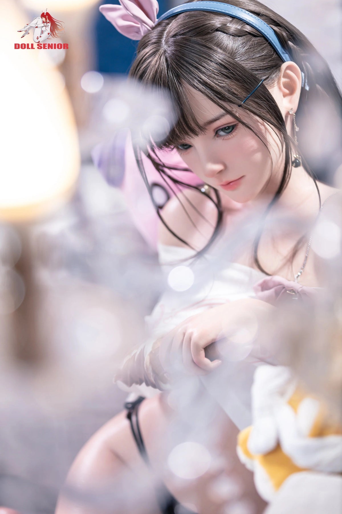 Realistic female doll with brown hair, blue headband, pink bunny ears, and soft skin texture wearing white top