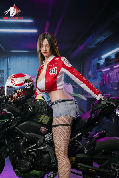 Realistic female doll in red racing jacket and denim shorts posing beside a black motorcycle in a neon-lit garage.
